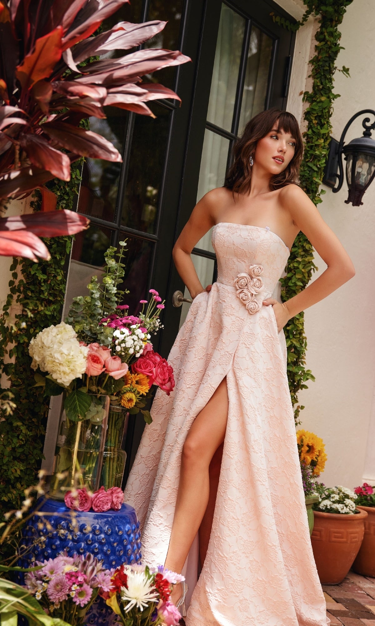 A woman in the Rosette A-Line Prom Dress, Ava Presley 40097—strapless with blush lace and a thigh-high slit—stands by a glass door amid vibrant potted flowers and lush greenery.